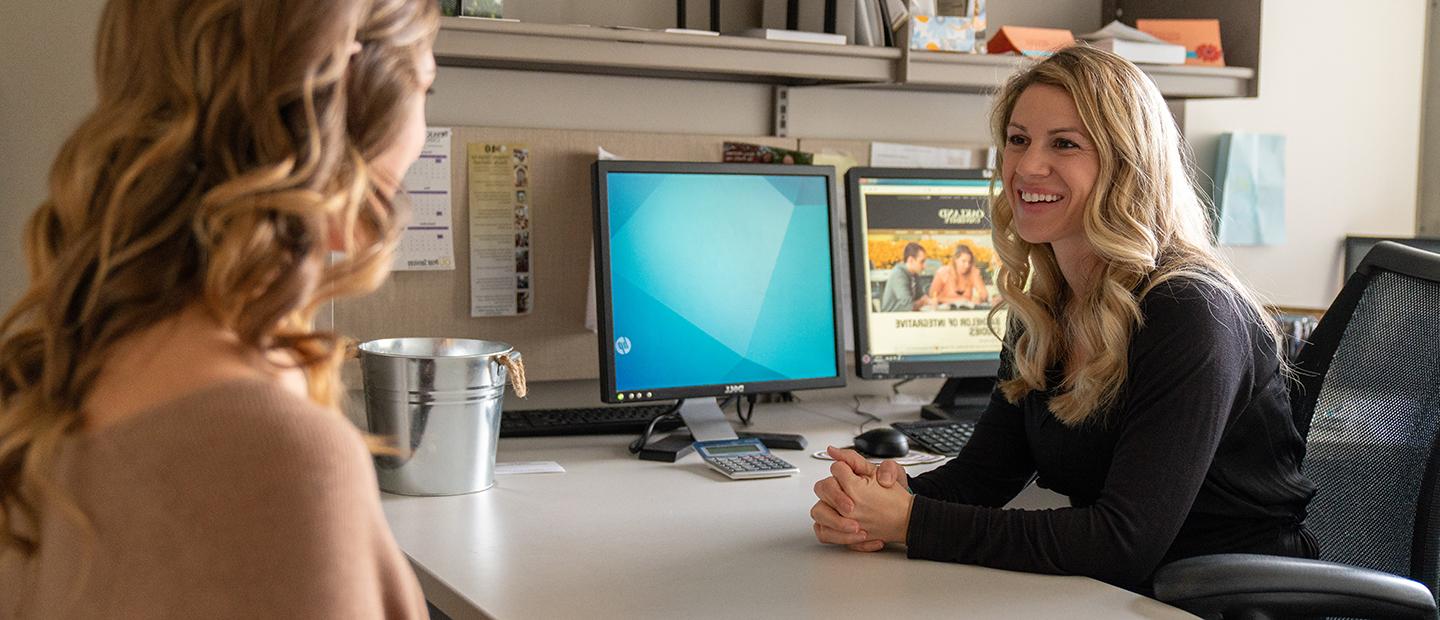 two women seated across from each other at a computer desk, talking and smiling.